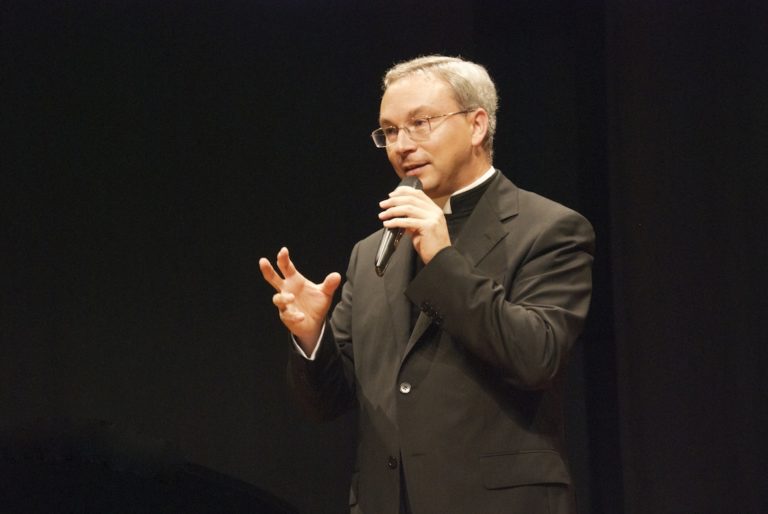 Don Alessio Geretti parla in pubblico con un microfono in mano, su fondo scuro.